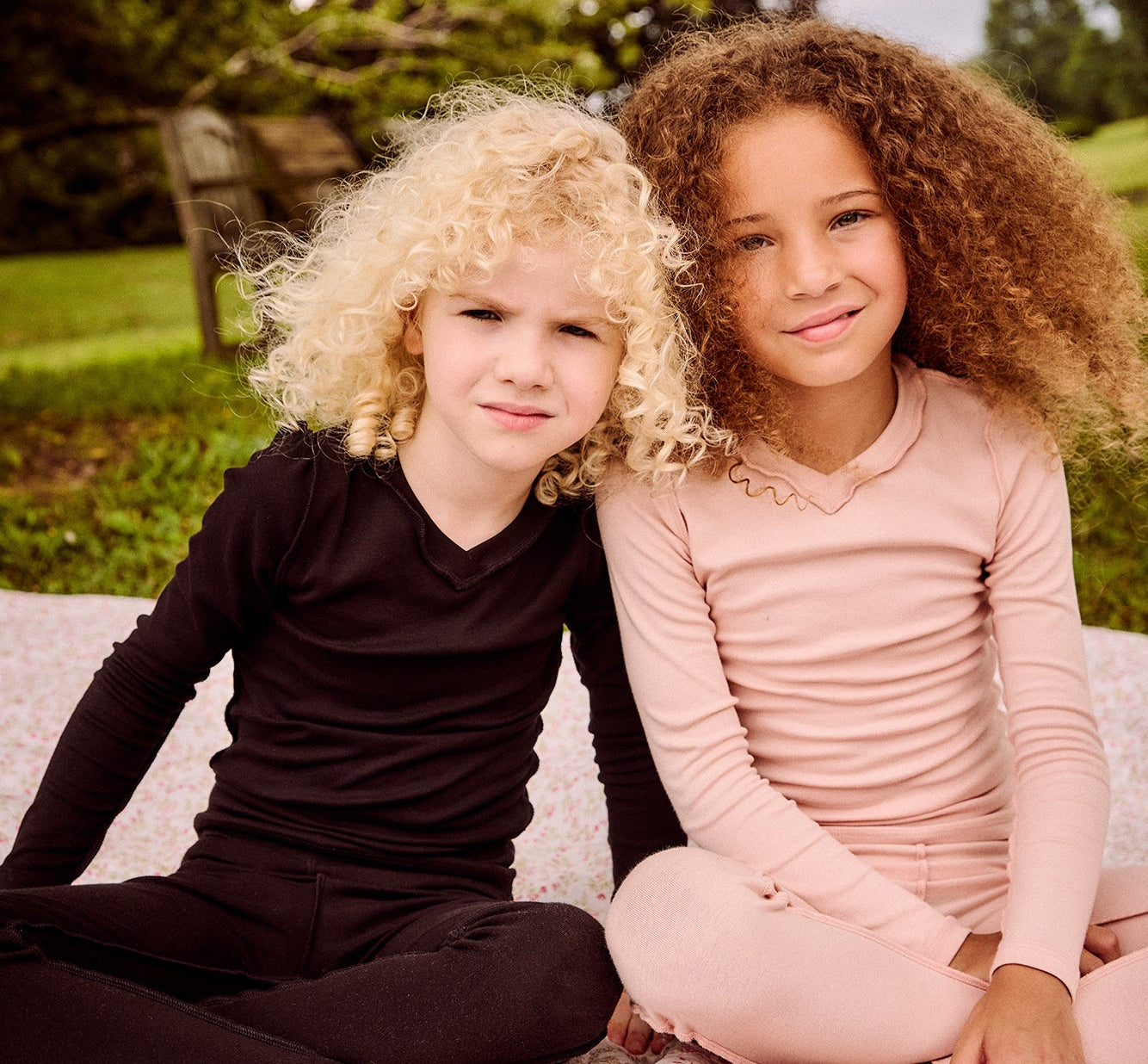Two young girls sitting side-by-side outdoors on a floral blanket, wearing matching long-sleeve activewear sets. The girl on the left wears a dark brownish-black set, and the girl on the right wears a light dusty pink set.
