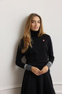 A woman with long blonde hair wearing a black turtleneck sweater with contrasting gray cuffs and a small red embroidered logo on the chest, posing against a light gray wall.