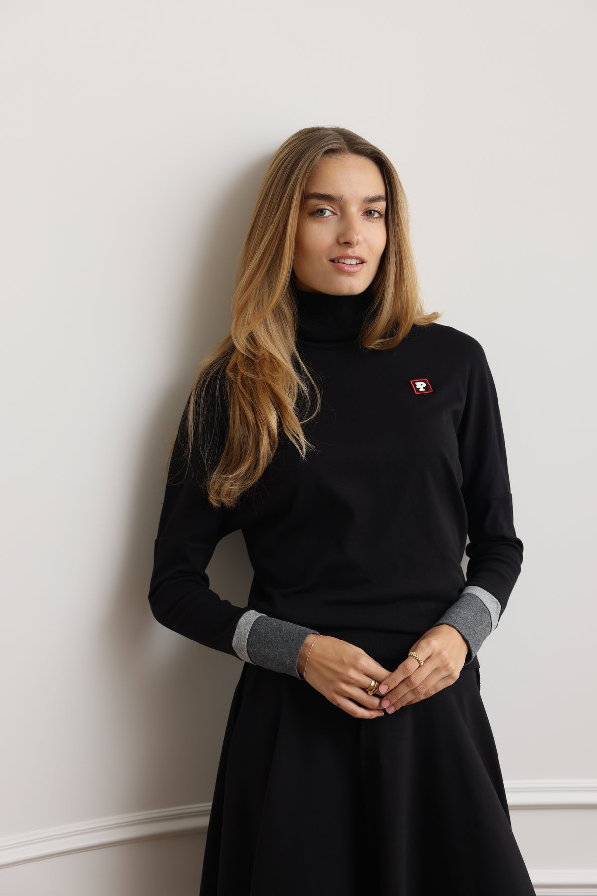 A woman with long blonde hair wearing a black turtleneck sweater with contrasting gray cuffs and a small red embroidered logo on the chest, posing against a light gray wall.