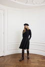 A woman stands in a white room with decorative wall molding and a wood parquet floor, wearing a black long-sleeve dress with a matching black beret, dark brown knee-high boots, and a small red lapel pin.