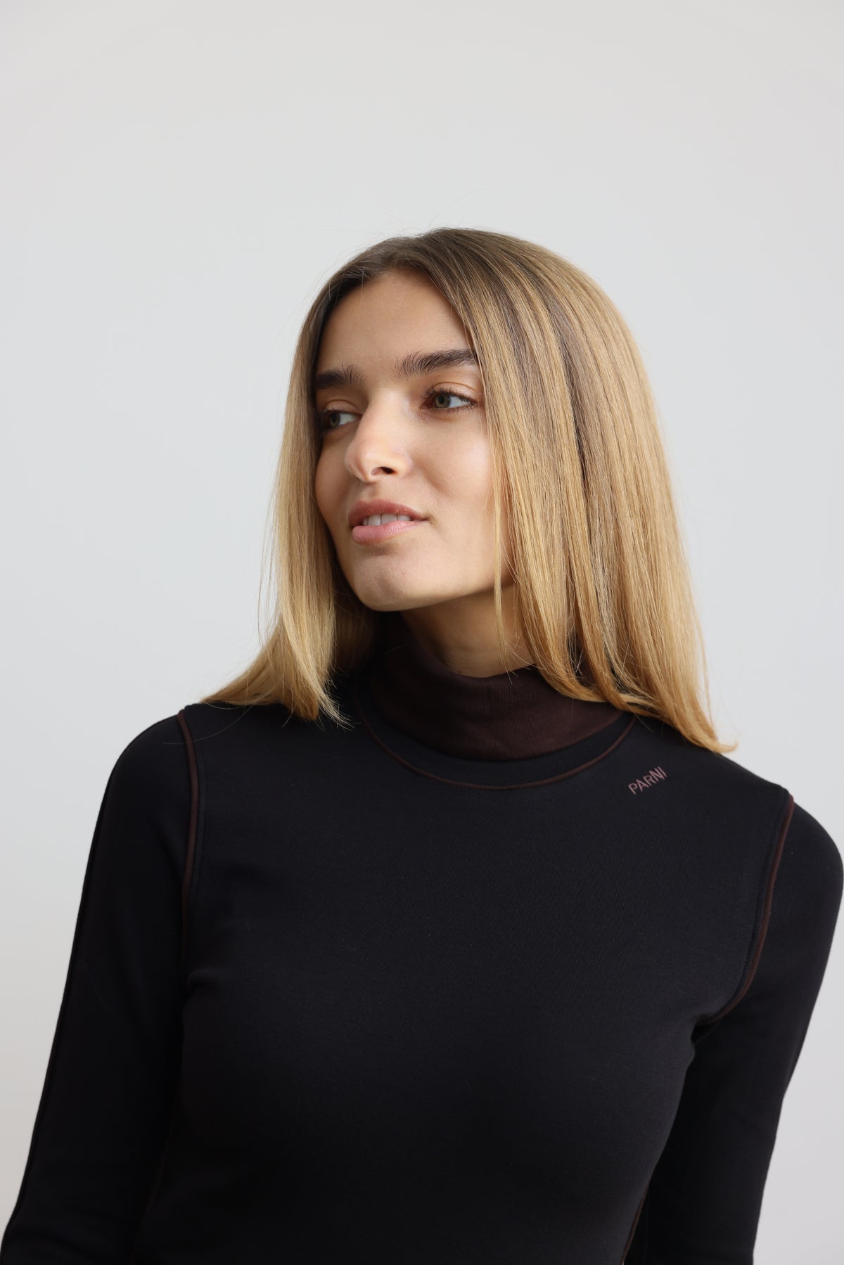 A woman with blonde hair wearing a black long-sleeve mock neck top with subtle brown contrast stitching, photographed against a light gray background.