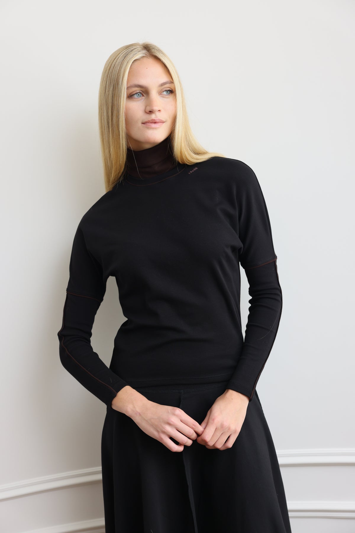 A blonde woman wearing a black long-sleeve mock neck top and black skirt, standing against a white wall.