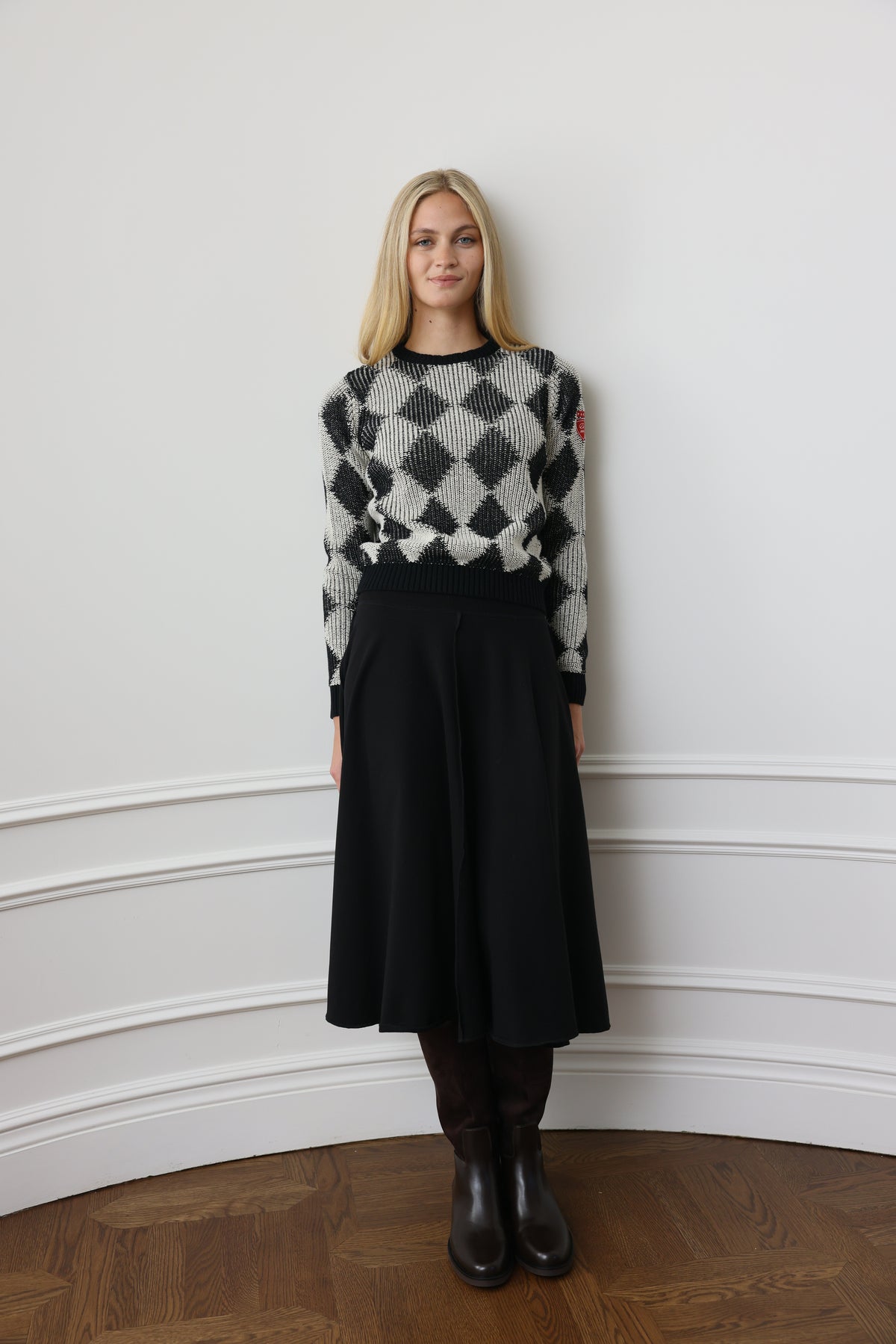 A woman wearing a black and white harlequin pattern knit sweater, a black skirt, and black boots, standing against a white wall with molding.