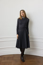 A woman stands in a room with white paneled walls and a wooden floor, wearing a dark charcoal gray long-sleeve mock-neck dress with a flared skirt, paired with dark tights and black ankle boots.