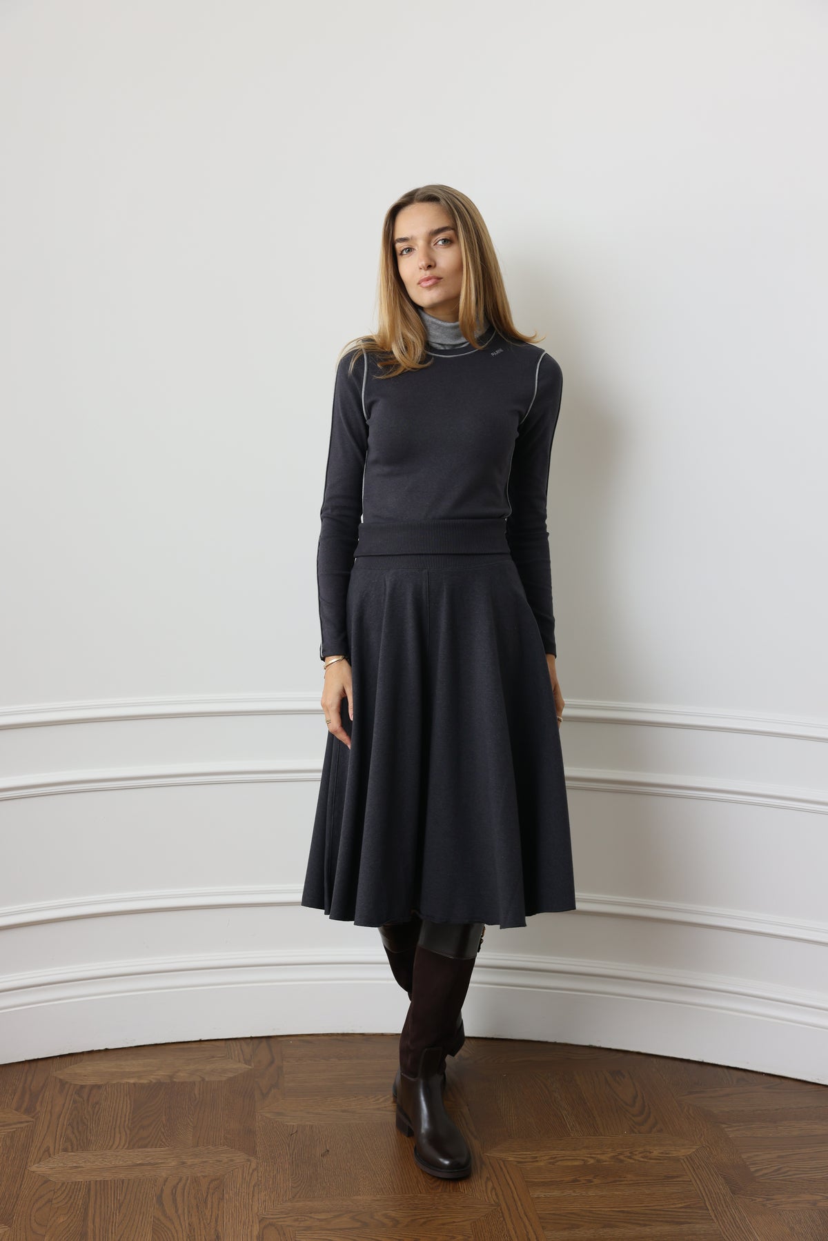 A woman stands in a room with white paneled walls and a wooden floor, wearing a dark charcoal gray long-sleeve mock-neck dress with a flared skirt, paired with dark tights and black ankle boots.