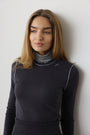 A woman with long light brown hair wearing a dark charcoal gray long-sleeve mock neck sweater with a gray turtleneck underneath, standing against a plain light gray wall.