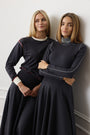 Two women posing in matching dark navy long-sleeve athletic tops and flared skirts against a light gray wall.