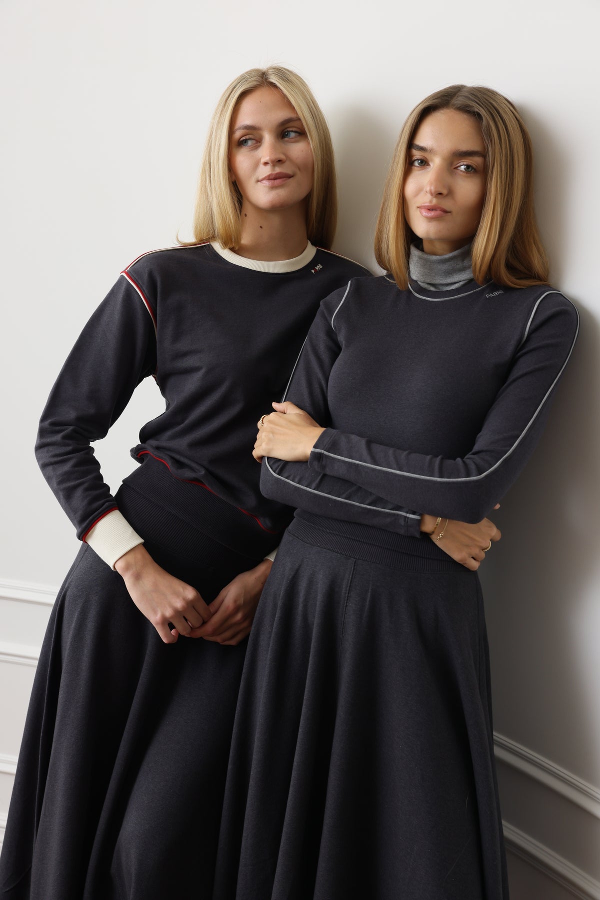 Two women posing in matching dark navy long-sleeve athletic tops and flared skirts against a light gray wall.
