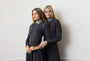 Two women modeling matching dark charcoal gray turtleneck sweaters and long skirts against a plain off-white wall.