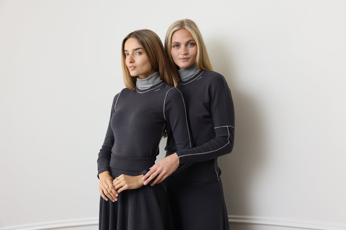 Two women modeling matching dark charcoal gray turtleneck sweaters and long skirts against a plain off-white wall.