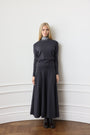 A woman stands against a white paneled wall wearing a dark charcoal gray knit mock-neck sweater and matching long skirt, paired with black ankle boots.