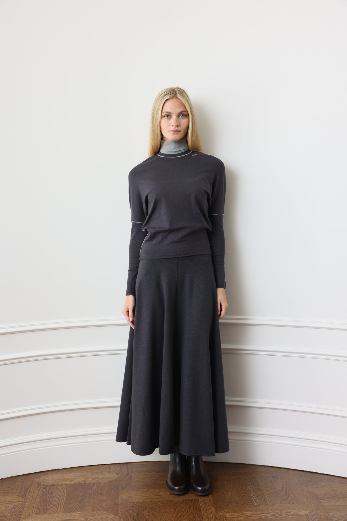 A woman stands against a white paneled wall wearing a dark charcoal gray knit mock-neck sweater and matching long skirt, paired with black ankle boots.