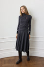 A woman wearing a dark charcoal gray long-sleeve midi dress with white stripe details on the sleeves and a small red embroidered logo on the chest, paired with black boots, standing in an indoor setting with a white wall and wooden parquet floor.