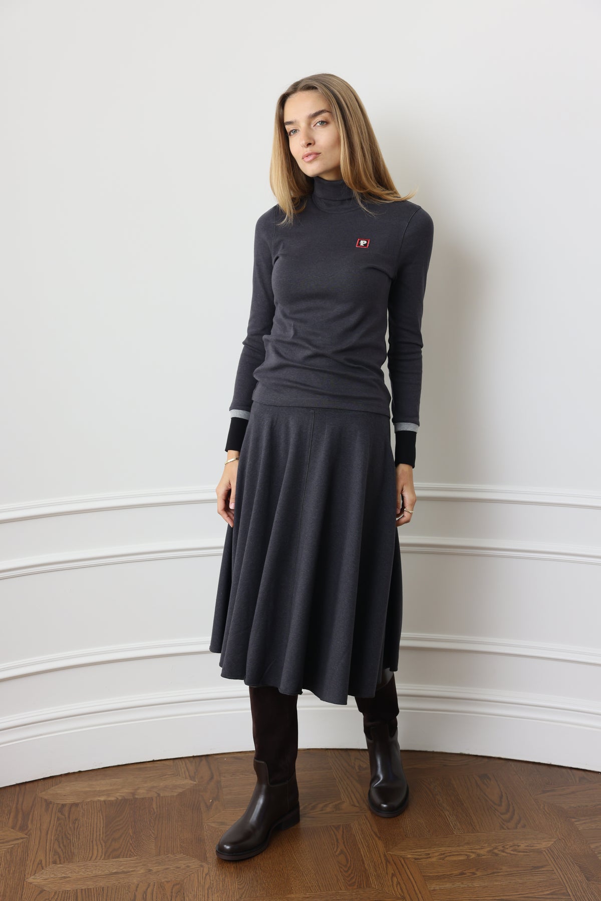 A woman wearing a dark charcoal gray long-sleeve midi dress with white stripe details on the sleeves and a small red embroidered logo on the chest, paired with black boots, standing in an indoor setting with a white wall and wooden parquet floor.