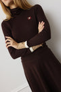 A woman wearing a dark brown long-sleeve mock-neck dress with a small red and white logo patch on the chest, posed against a light gray wall.