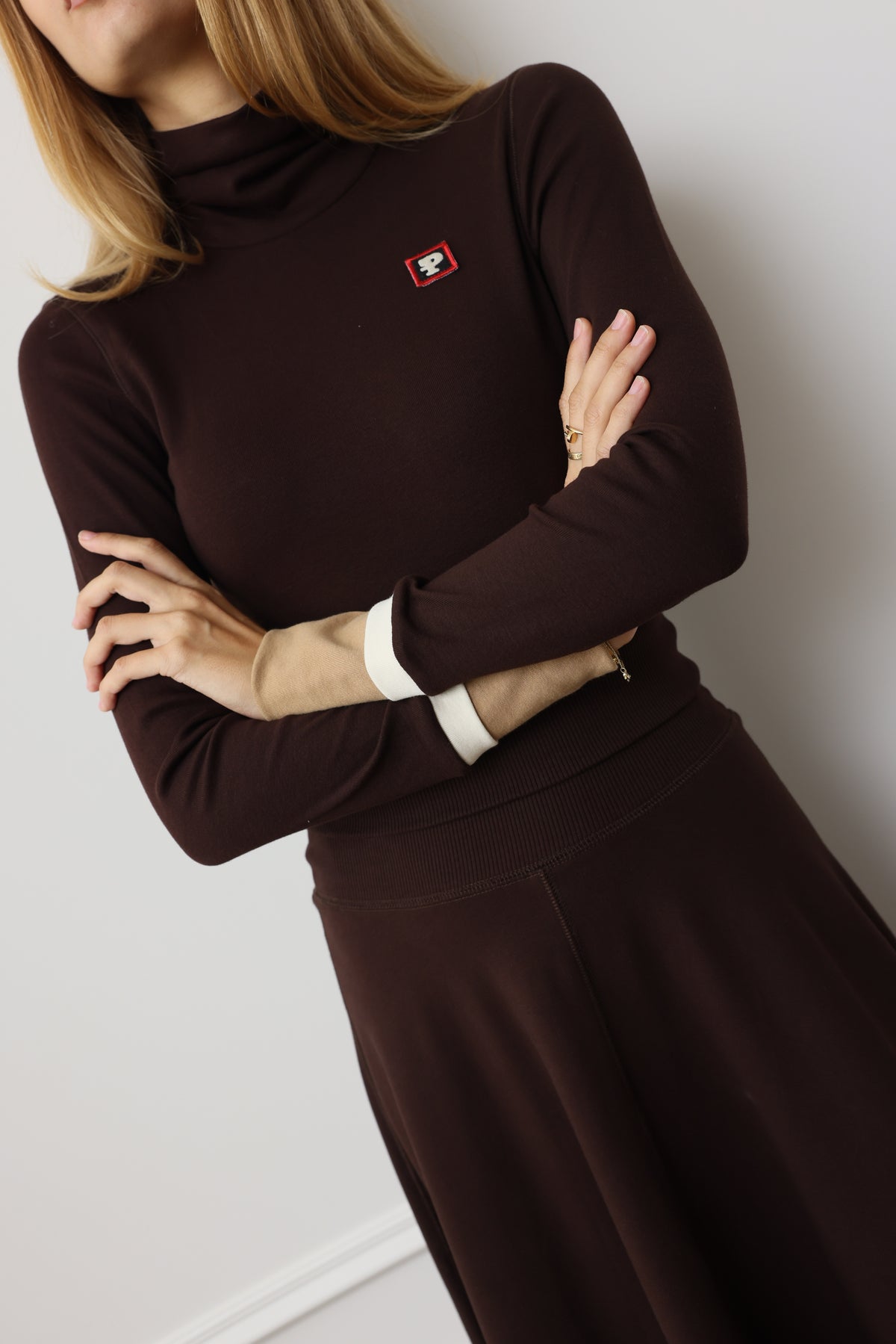 A woman wearing a dark brown long-sleeve mock-neck dress with a small red and white logo patch on the chest, posed against a light gray wall.