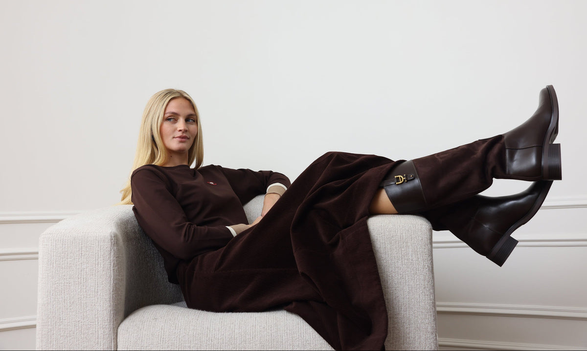 A woman reclining on a light gray armchair, wearing a dark brown long-sleeve dress and matching dark brown leather knee-high boots with a small gold logo detail.