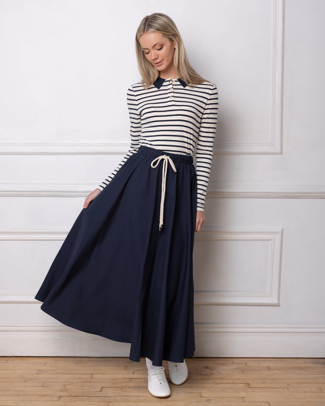 Model wearing navy maxi skirt and striped polo standing against a white wall.