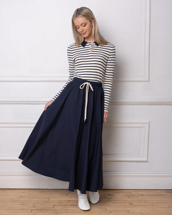 Model wearing navy maxi skirt and striped polo standing against a white wall.