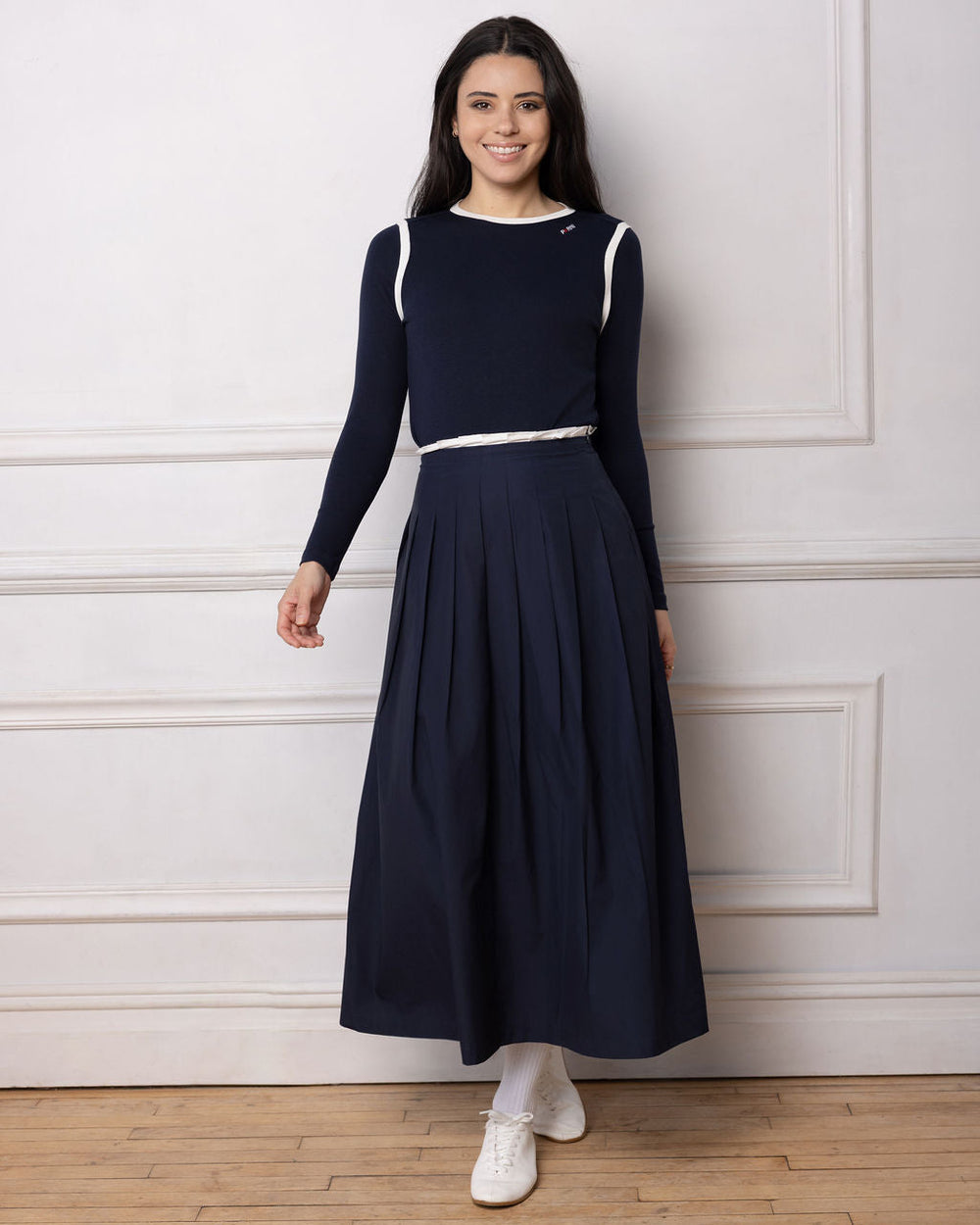 Model wearing a navy pleated skirt and navy top standing against a white wall.