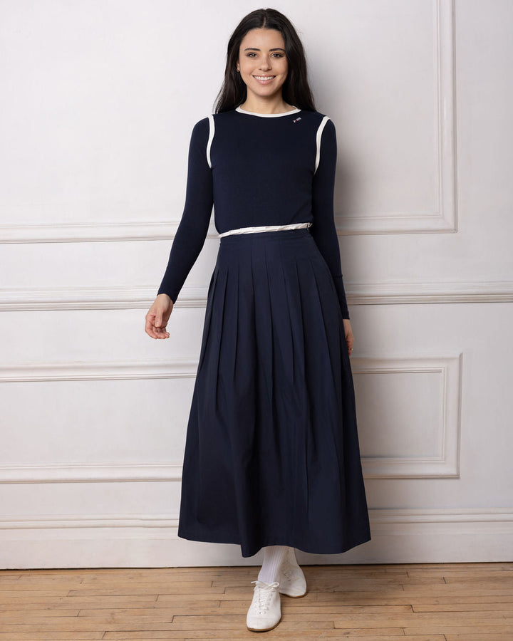 Model wearing a navy pleated skirt and navy top standing against a white wall.
