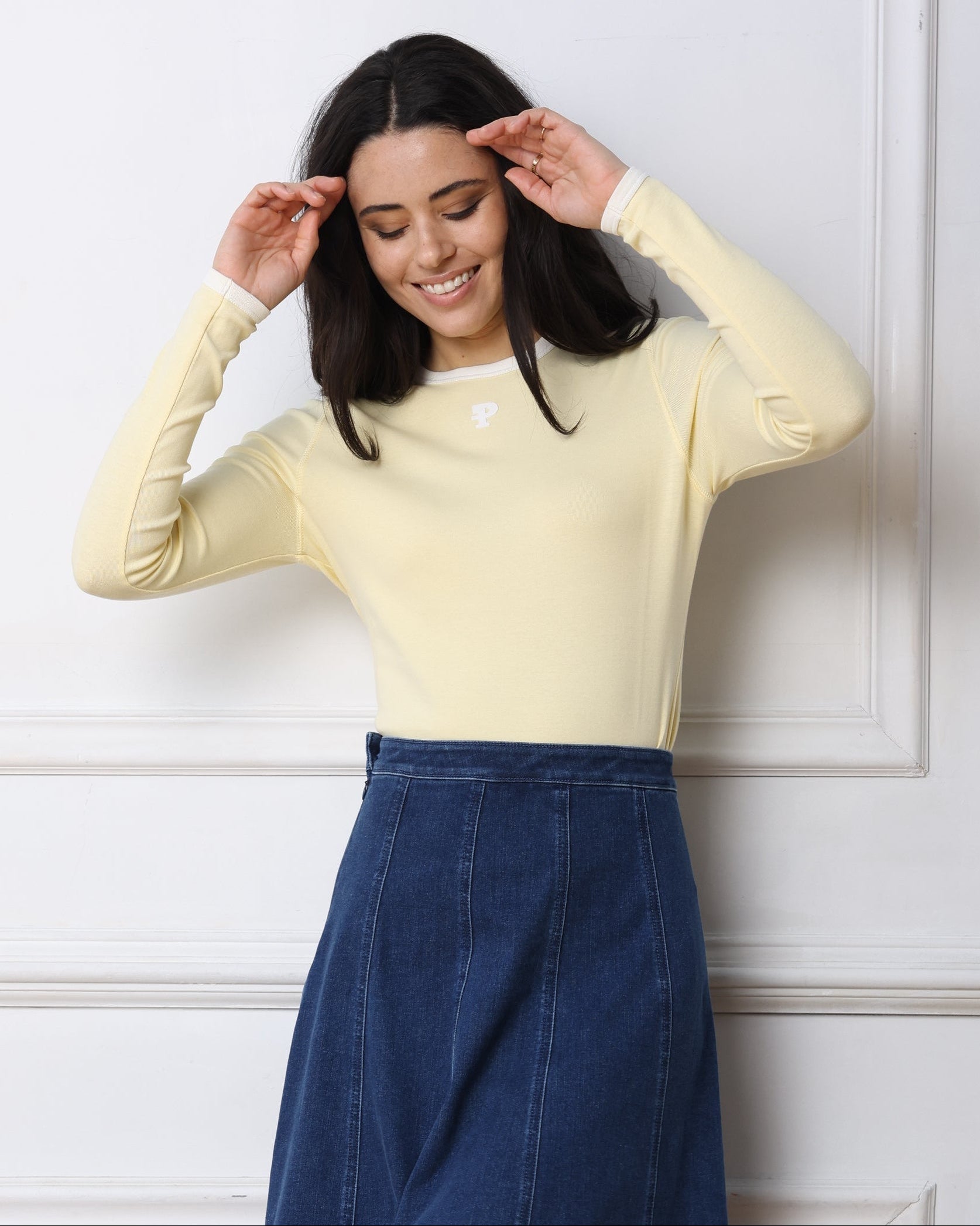 Woman wearing a light yellow sweater and blue skirt against a white wall
