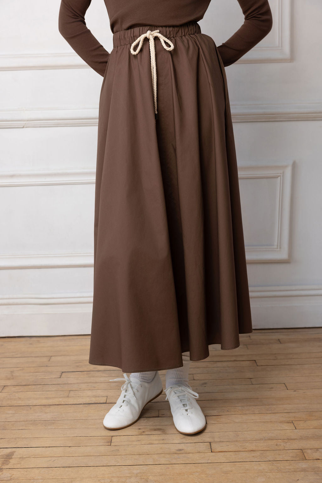 Close up of Model wearing brown maxi with rope belt on  standing near a white wall.