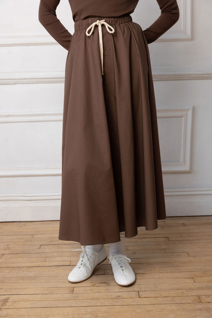 Close up of Model wearing brown maxi with rope belt on  standing near a white wall.