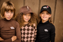 Three children wearing matching custom apparel from 'Crazy Dog Outfitters' posed together against a brown wood-paneled wall. The child on the left wears a brown turtleneck, the center child wears a brown and pink argyle shirt with a brown cap, and the child on the right wears a black turtleneck and black cap.