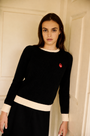 A woman wearing a black ribbed knit sweater with cream ribbed trim at the crew neck, cuffs, and hem, featuring a small red embroidered logo on the left chest, posed against a cream-colored paneled door.