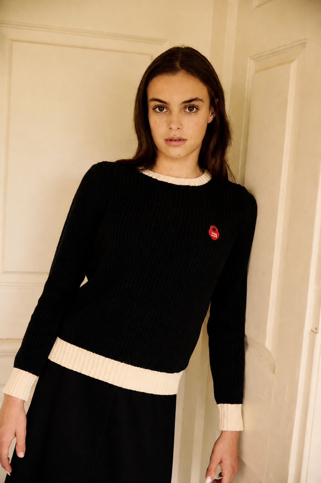 A woman wearing a black ribbed knit sweater with cream ribbed trim at the crew neck, cuffs, and hem, featuring a small red embroidered logo on the left chest, posed against a cream-colored paneled door.
