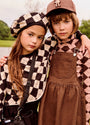 Two children posing outdoors in coordinated autumn fashion. The girl wears a black and beige checkered beret and matching checkered top, while the boy wears a brown cap, a brown corduroy overalls dress, and a brown-and-pink argyle shirt.