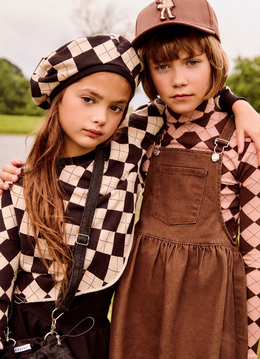 Two children posing outdoors in coordinated autumn fashion. The girl wears a black and beige checkered beret and matching checkered top, while the boy wears a brown cap, a brown corduroy overalls dress, and a brown-and-pink argyle shirt.