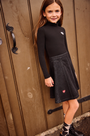 A young girl with long brown hair stands in front of a rustic wooden door, wearing a black long-sleeve mock neck top and a black denim A-line skirt with small red heart details.