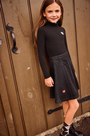 A young girl with long brown hair stands in front of a rustic wooden door, wearing a black long-sleeve mock neck top and a black denim A-line skirt with small red heart details.