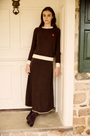 A woman stands in a doorway wearing a dark brown ribbed knit sweater and matching long skirt with cream trim, paired with dark brown boots.