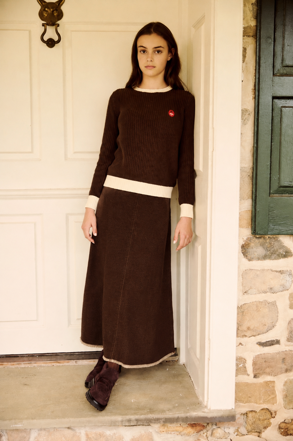 A woman stands in a doorway wearing a dark brown ribbed knit sweater and matching long skirt with cream trim, paired with dark brown boots.