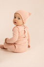 A baby wearing a soft dusty pink long-sleeve footed romper with a matching pointed hat, sitting against a solid pale beige studio background.