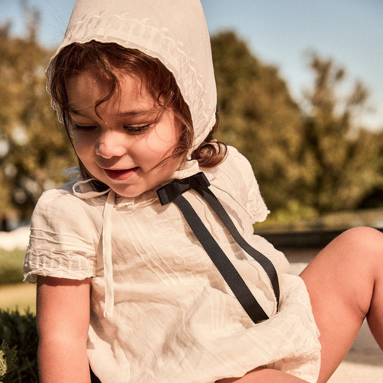 Child wearing a white hooded outfit with a black bow sitting outdoors.