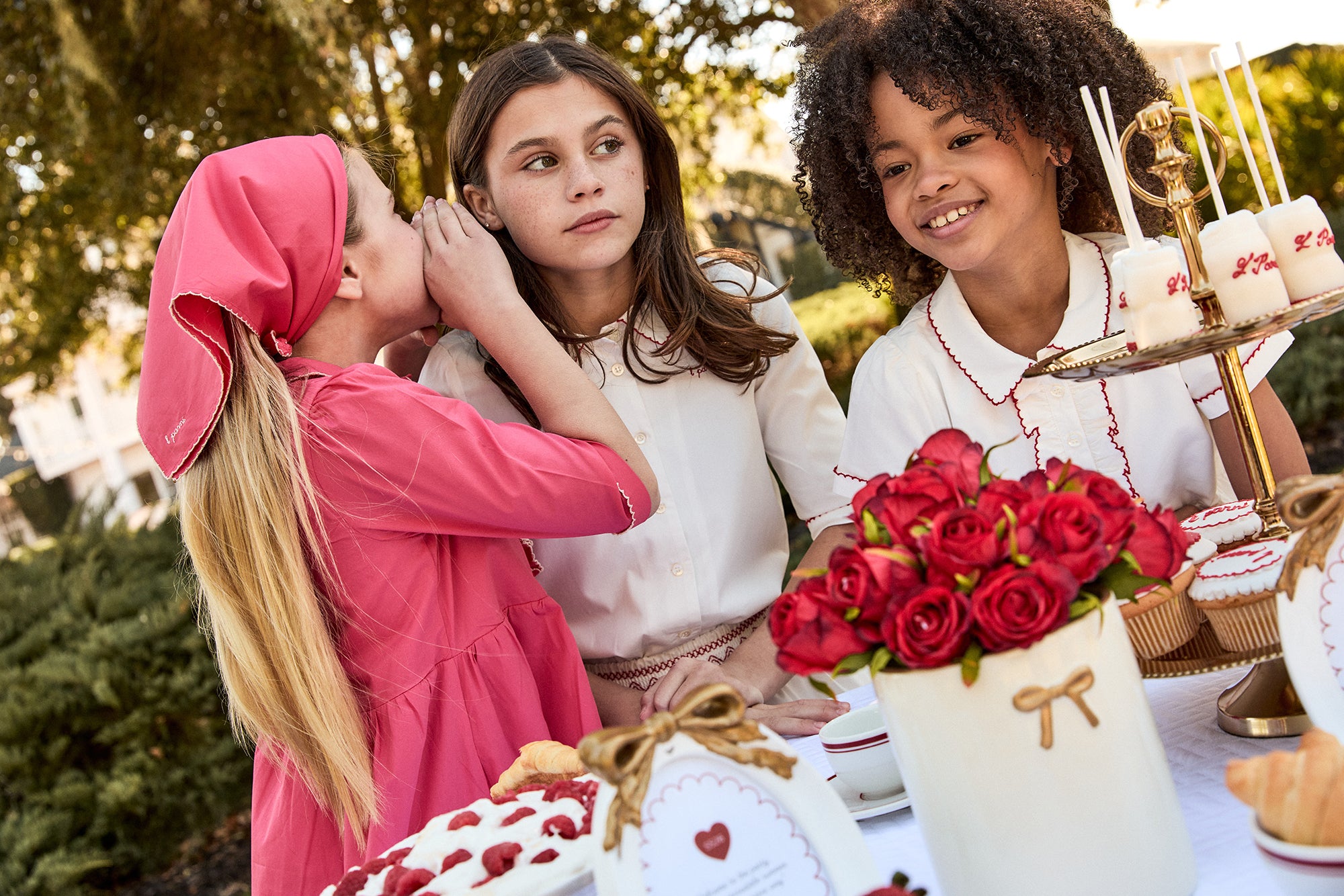 Three young girls at a table with a cake and flowers, outdoors.