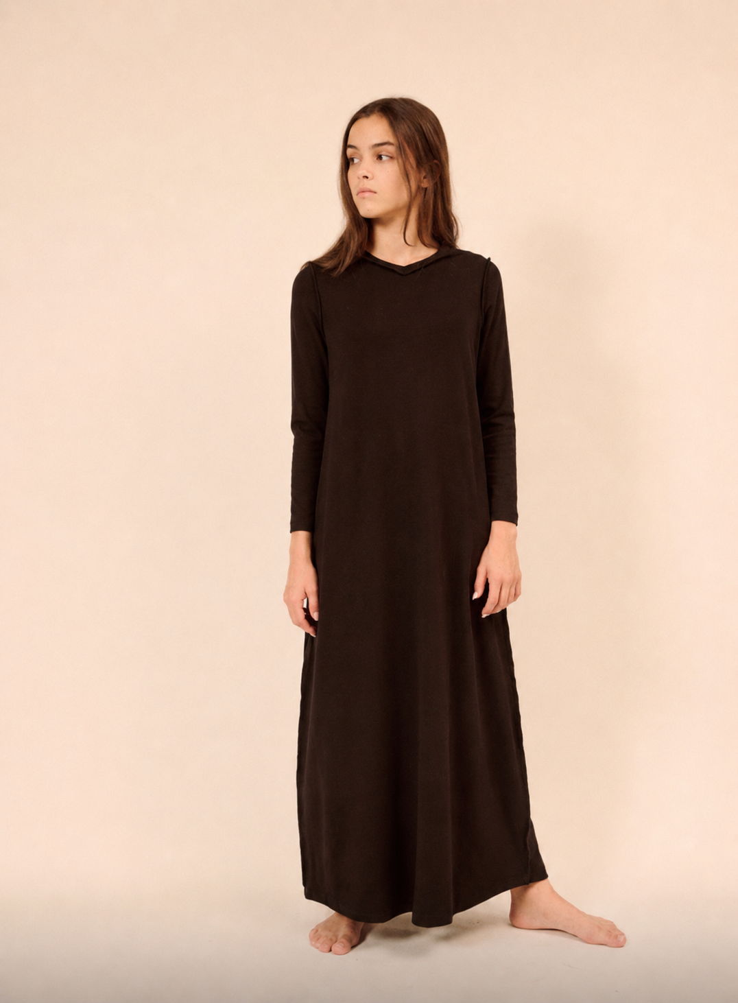 A woman modeling a long-sleeved, ankle-length dark brown nightgown  against a solid beige studio background.