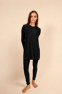A woman modeling a black long-sleeve top and black legging pajamas, standing against a solid beige studio background.
