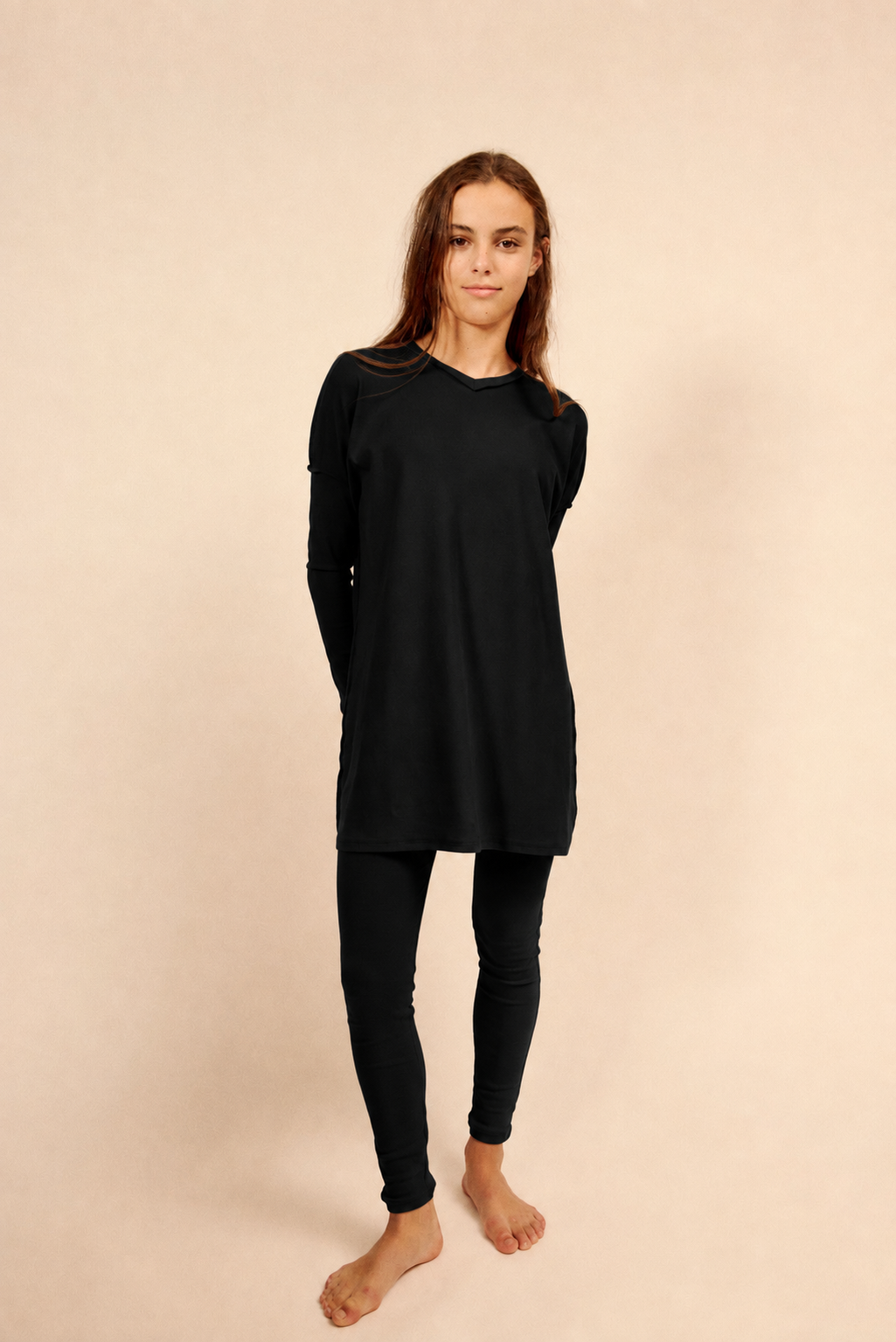 A woman modeling a black long-sleeve top and black legging pajamas, standing against a solid beige studio background.