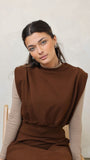 Woman wearing a brown dress with a beige long-sleeve shirt underneath, sitting on a chair against a plain background.