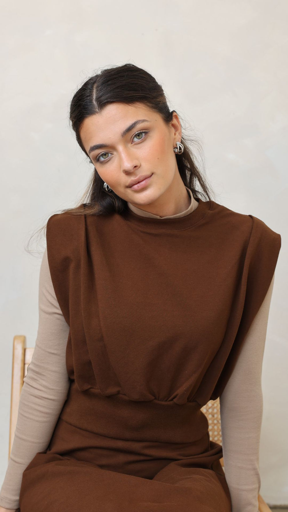 Woman wearing a brown dress with a beige long-sleeve shirt underneath, sitting on a chair against a plain background.