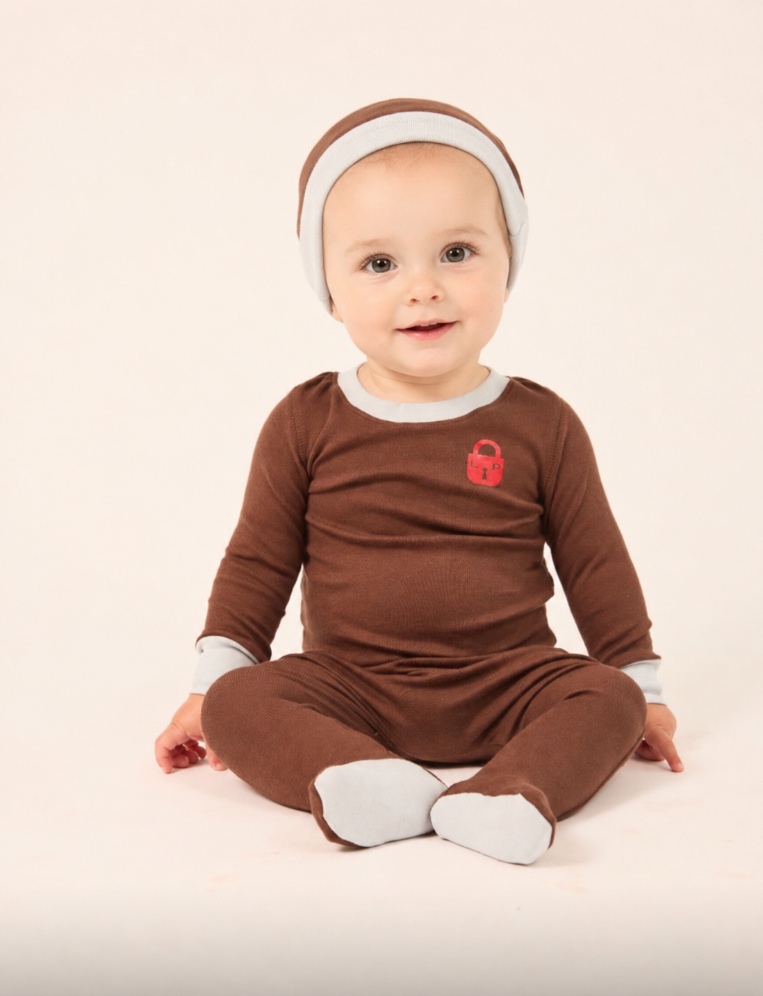 A baby wearing a brown and white footed jumpsuit and matching headband, sitting against a plain light background.