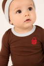 A close-up of a baby wearing a brown long-sleeve shirt with a light gray ribbed collar and a small red padlock graphic on the chest, paired with a matching brown and light gray beanie against a plain light background.