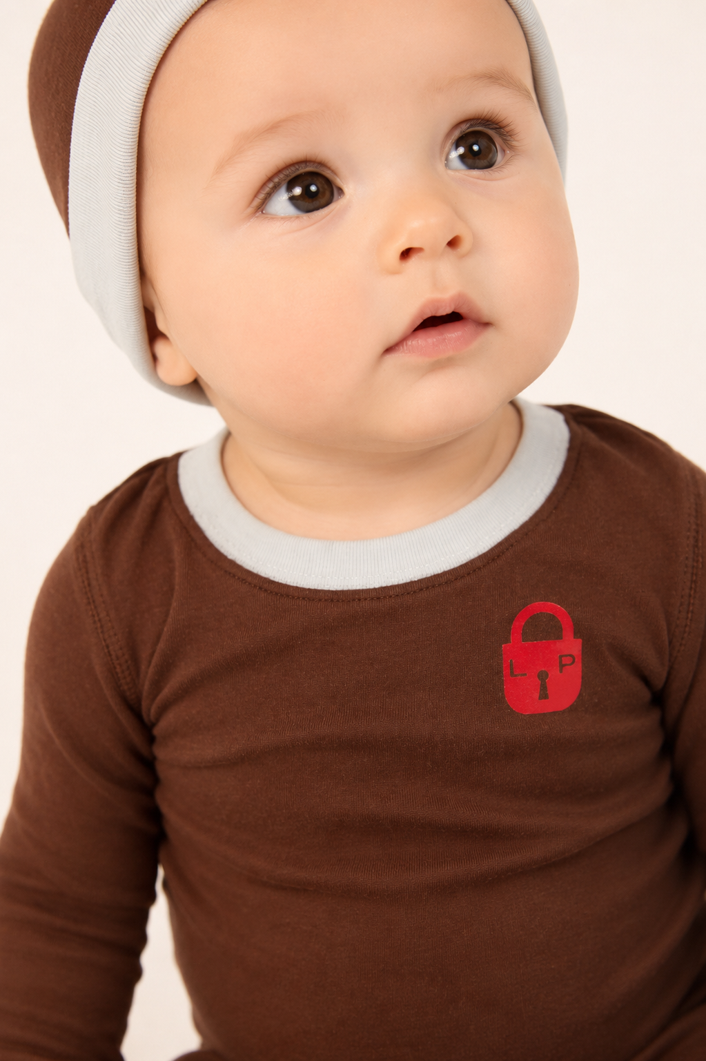 A close-up of a baby wearing a brown long-sleeve shirt with a light gray ribbed collar and a small red padlock graphic on the chest, paired with a matching brown and light gray beanie against a plain light background.