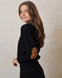 Woman wearing a black outfit with brown elbow patches against a plain background