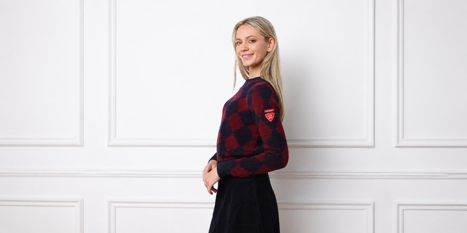 A woman with blonde hair stands in profile against a white paneled wall, wearing a red and black checkered long-sleeve sweater with a small red shield logo on the sleeve, paired with a dark skirt.